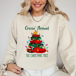 May include: A cream-colored sweatshirt features a Christmas tree design made of red and green Crocs, adorned with string lights and a gold star. The text "Crocin' Around THE CHRISTMAS TREE" is printed above and below the tree.