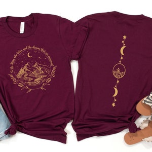 Op de afbeelding: Bordeauxrode t-shirts met gouden grafische ontwerpen. E&eacute;n shirt heeft een berg- en maanontwerp met de tekst "To the stars who listen and the dreams that are answered". Het andere shirt heeft een verticaal maan- en sterrenontwerp.