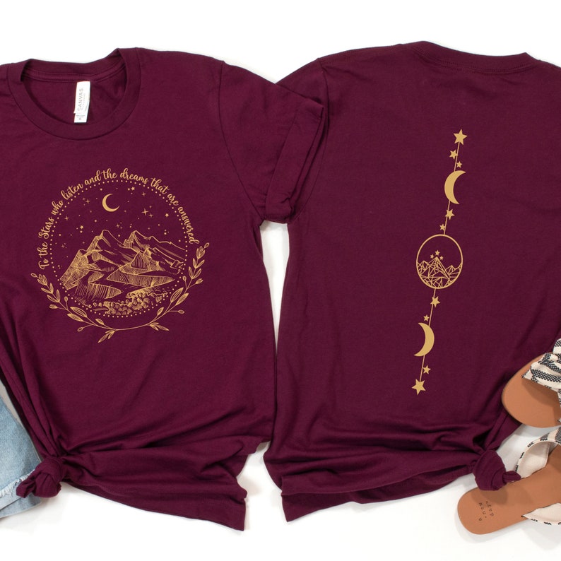 Op de afbeelding: Bordeauxrode t-shirts met gouden grafische ontwerpen. E&eacute;n shirt heeft een berg- en maanontwerp met de tekst "To the stars who listen and the dreams that are answered". Het andere shirt heeft een verticaal maan- en sterrenontwerp.