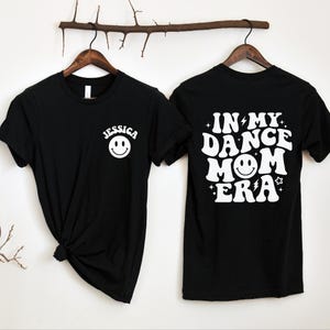 Op de afbeelding: Zwart T-shirt met witte tekst die "In My Dance Mom Era" zegt met een smiley. De voorkant van het shirt heeft een witte smiley en de naam "Jessica" erboven.