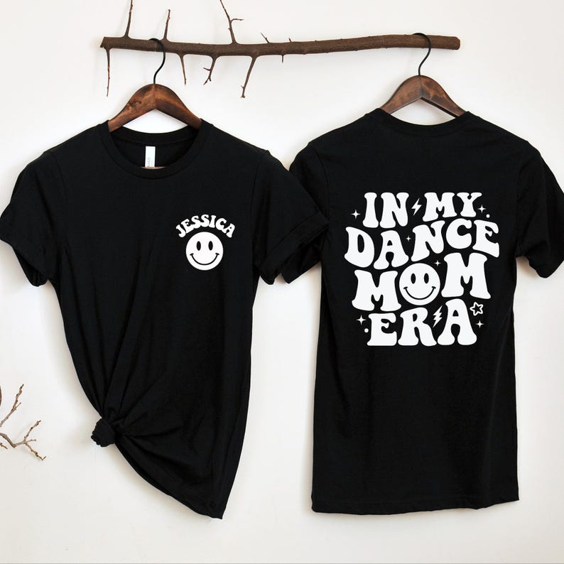 Op de afbeelding: Zwart T-shirt met witte tekst die "In My Dance Mom Era" zegt met een smiley. De voorkant van het shirt heeft een witte smiley en de naam "Jessica" erboven.