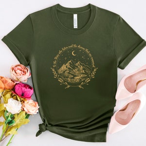 Op de afbeelding: Olijfgroen t-shirt met een gouden afbeelding van bergen, een maan en sterren, omcirkeld door een krans. De tekst luidt: "To the stars who listen and the dreams that are answered."