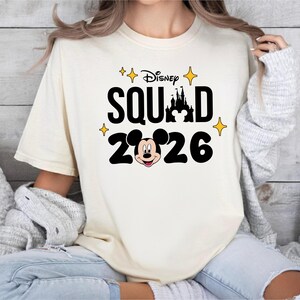 A woman wearing a white t-shirt with a Disney Mickey Mouse graphic and the text "Disney SQUAD 2026" printed on it.