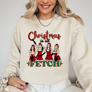 May include: Beige sweatshirt with the text "Christmas is so FETCH" in red and green. The graphic features four women in Santa outfits. The sweatshirt is a crew neck style.