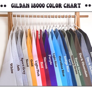 Op de afbeelding: Een kleurenkaart van Gildan 18000 sweatshirts die aan een houten rek hangen. De sweatshirts zijn in verschillende kleuren, waaronder wit, sportgrijs, zand, goud, lichtroze, rood, bordeauxrood, paars, lichtblauw en zwart. De tekst "GILDAN 18000 COLOR CHART" staat bovenaan.