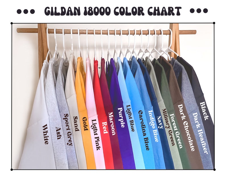 Op de afbeelding: Een kleurenkaart van Gildan 18000 sweatshirts die aan een houten rek hangen. De sweatshirts zijn in verschillende kleuren, waaronder wit, sportgrijs, zand, goud, lichtroze, rood, bordeauxrood, paars, lichtblauw en zwart. De tekst "GILDAN 18000 COLOR CHART" staat bovenaan.