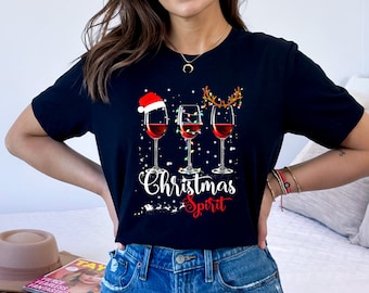 May include: Black t-shirt with a Christmas-themed graphic. The design features three wine glasses filled with red liquid, each adorned with festive elements: a Santa hat, string lights, and reindeer antlers. The words "Christmas Spirit" are written below.