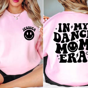 Op de afbeelding: Roze sweatshirt met zwarte tekst die "In My Dance Mom Era" zegt met een smiley in het midden. De voorkant van het sweatshirt heeft een zwarte smiley en de naam "Jessica" erboven.