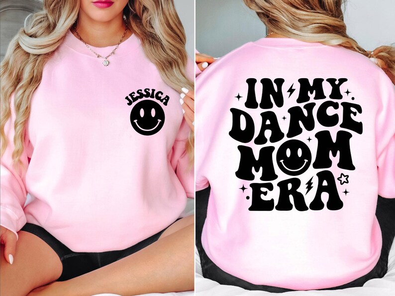 Op de afbeelding: Roze sweatshirt met zwarte tekst die "In My Dance Mom Era" zegt met een smiley in het midden. De voorkant van het sweatshirt heeft een zwarte smiley en de naam "Jessica" erboven.