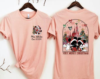 May include: Two light pink t-shirts hanging on a wooden hanger. The front of the shirt has a graphic of Mickey and Minnie Mouse in Santa hats with the text "Mrs. William Est 2024". The back of the shirt has a graphic of Mickey and Minnie Mouse in front of a castle with the text "VERY MERRY CHRISTMAS".