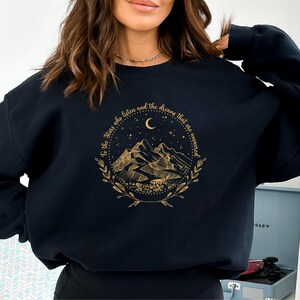 Op de afbeelding: Een marineblauwe sweatshirt met een gouden grafisch ontwerp met bergen, een halve maan en sterren. De tekst luidt: "To the Stars who listen and the dreams that are answered." Het ontwerp is ingesloten in een cirkelvormige krans.