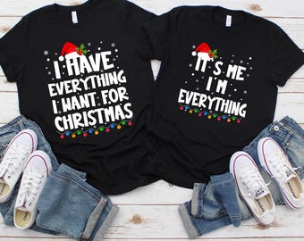 Pode incluir: Duas camisetas pretas com texto e gráficos natalinos. Uma diz "I HAVE EVERYTHING I WANT FOR CHRISTMAS", a outra "IT'S ME I'M EVERYTHING". Ambas apresentam gorros de Papai Noel e luzes.