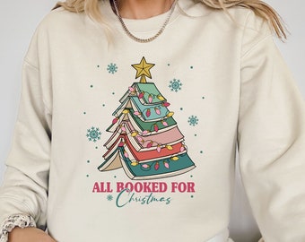 May include: Cream-colored sweatshirt featuring a Christmas tree design made of books. The tree is adorned with string lights and topped with a gold star. The text "ALL BOOKED FOR Christmas" is printed below the tree in red and teal.
