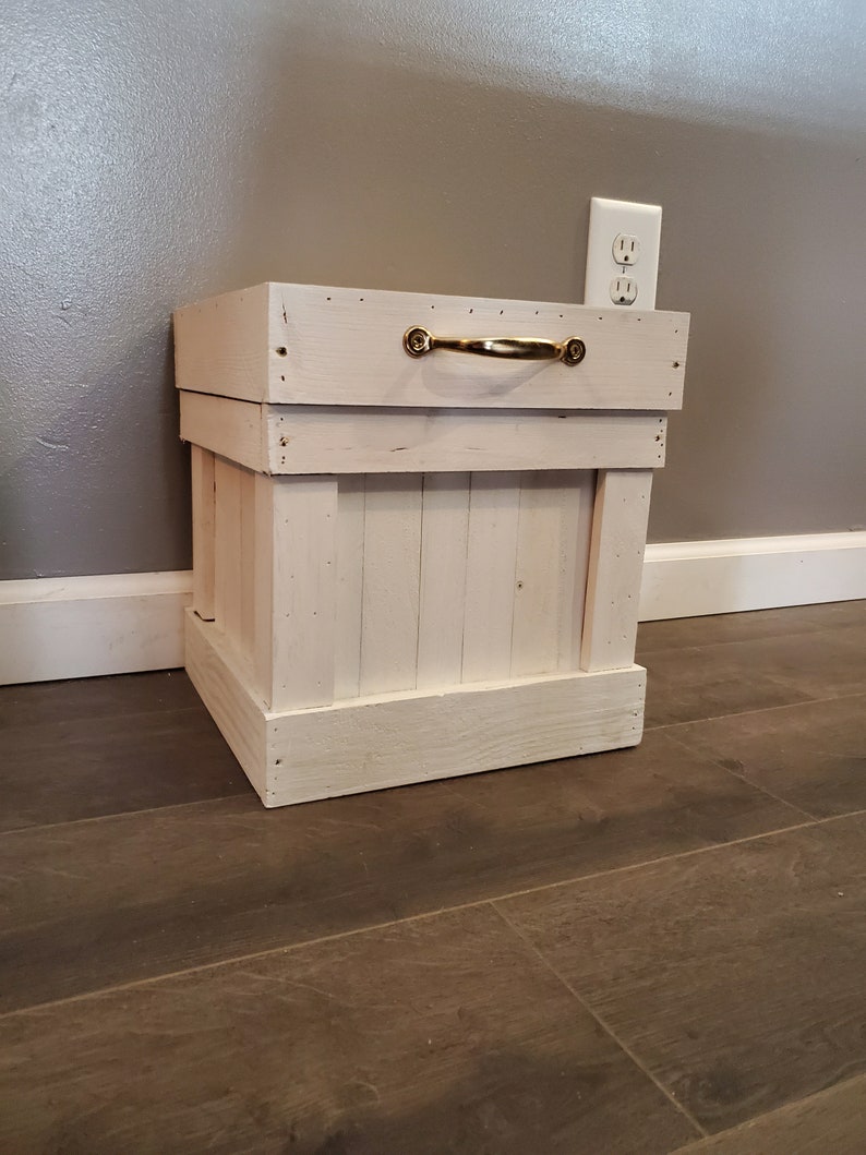 May include: White wooden storage box with a gold metal handle. The box is sitting on a wood floor.