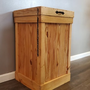 May include: A wooden trash can with a hinged lid and a black metal handle. The lid is held closed with a black chain.