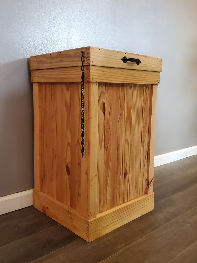 May include: A wooden trash can with a hinged lid and a black metal handle. The lid is held closed with a black chain.
