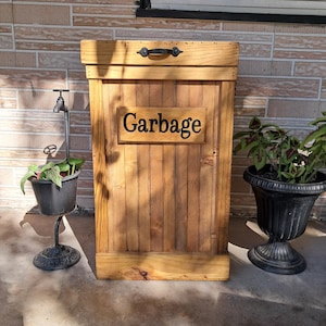 May include: A wooden garbage can with a hinged lid and a black metal handle. The can is painted a light brown color and has a sign that says "Garbage" on the front.