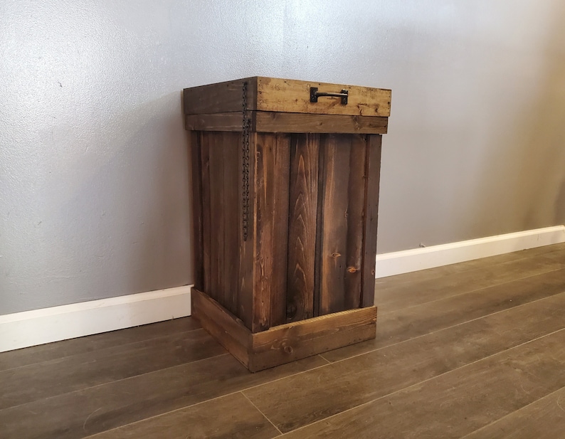 May include: A rustic wooden trash can with a hinged lid and a metal handle. The can is made of reclaimed wood and has a dark brown finish.
