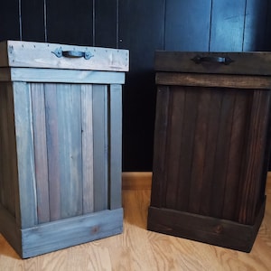 Pu&ograve; includere: Due bidoni della spazzatura in legno con coperchi e maniglie in metallo nero. Il bidone a sinistra &egrave; dipinto di un colore grigio chiaro, mentre il bidone a destra &egrave; dipinto di un colore marrone scuro.