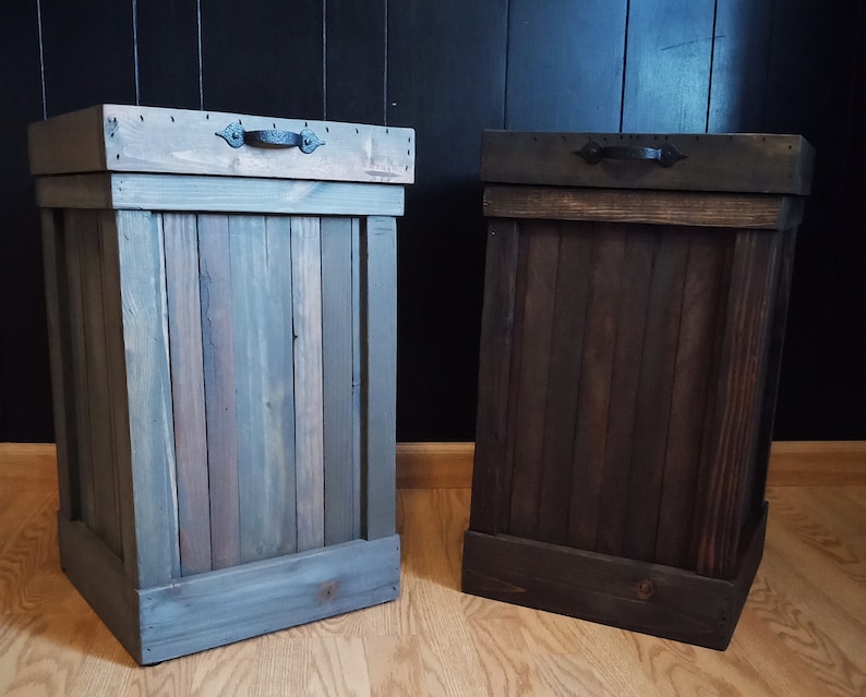 Pu&ograve; includere: Due bidoni della spazzatura in legno con coperchi e maniglie in metallo nero. Il bidone a sinistra &egrave; dipinto di un colore grigio chiaro, mentre il bidone a destra &egrave; dipinto di un colore marrone scuro.