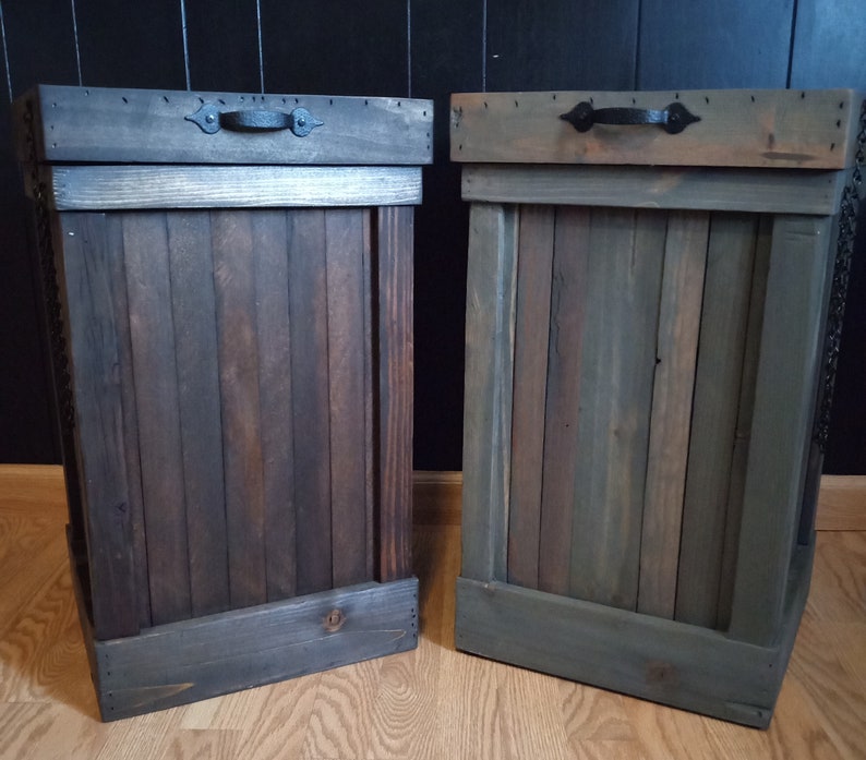 Pu&ograve; includere: Due bidoni della spazzatura in legno con maniglie in metallo nero. I bidoni della spazzatura sono realizzati in legno riciclato e hanno una finitura rustica. Un bidone della spazzatura &egrave; marrone scuro e l'altro &egrave; grigio-verde chiaro.
