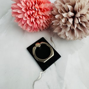 May include: A black phone ring holder with a silver metal ring. The ring holder is on a white marble surface.