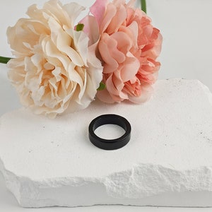 Op de afbeelding: Een zwarte ring op een witte steen met twee roze en witte bloemen erachter.