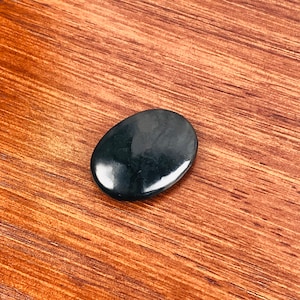 Puede incluir: Una piedra de obsidiana negra lisa de forma ovalada. La piedra tiene una superficie pulida y un color oscuro, casi negro.