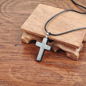 May include: A gray stone cross pendant on a black cord necklace. The cross is displayed on a small wooden stand with a dark brown finish. The background is a wooden surface with a natural wood grain.