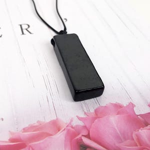 Może przedstawiać: Naszyjnik z czarną, prostokątną zawieszką i czarnym sznurkiem. Zawieszka jest gładka i błyszcząca. Tło stanowi biała drewniana powierzchnia z różowymi płatkami róż.
