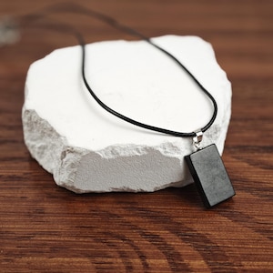 May include: A black rectangular pendant necklace with a silver clasp, hanging on a black cord, displayed on a white rock.