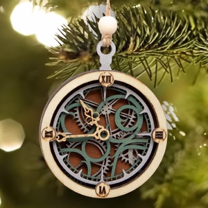 May include: Wooden Christmas ornament in the shape of a clock. The clock has a layered design with gears and Roman numerals. The ornament is suspended by a string and a wooden bead. The clock face is brown, green, and beige.