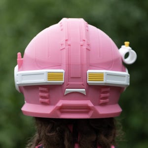 Pu&ograve; includere: Un casco rosa dal design strutturato con una fascia orizzontale bianca al centro. La fascia presenta accenti rettangolari gialli su entrambi i lati. Un piccolo attacco circolare bianco con la parte superiore gialla &egrave; visibile sul lato destro.