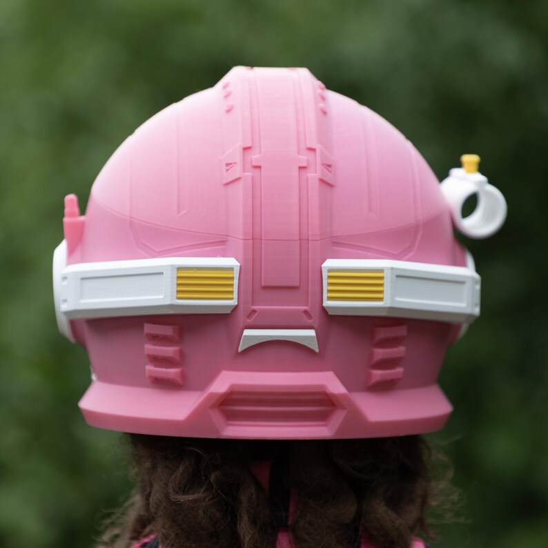 Pu&ograve; includere: Un casco rosa dal design strutturato con una fascia orizzontale bianca al centro. La fascia presenta accenti rettangolari gialli su entrambi i lati. Un piccolo attacco circolare bianco con la parte superiore gialla &egrave; visibile sul lato destro.