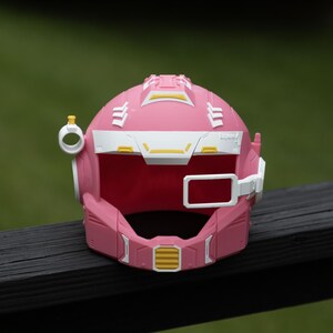 Pu&ograve; includere: Un casco in stile fantascienza di colore rosa, bianco e giallo, caratterizzato da un'area visiera prominente, un piccolo attacco laterale circolare con una parte superiore gialla e un telaio rettangolare laterale.