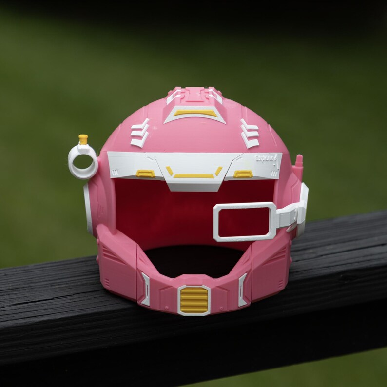 Pu&ograve; includere: Un casco in stile fantascienza di colore rosa, bianco e giallo, caratterizzato da un'area visiera prominente, un piccolo attacco laterale circolare con una parte superiore gialla e un telaio rettangolare laterale.