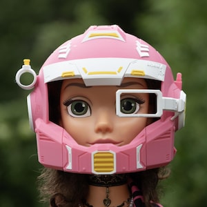 Pu&ograve; includere: Un casco futuristico rosa e bianco con accenti gialli indossato da una bambola. Il casco presenta un oculare rettangolare sopra l'occhio sinistro, un attacco circolare sul lato destro e un dettaglio di ventilazione giallo sulla mentoniera.