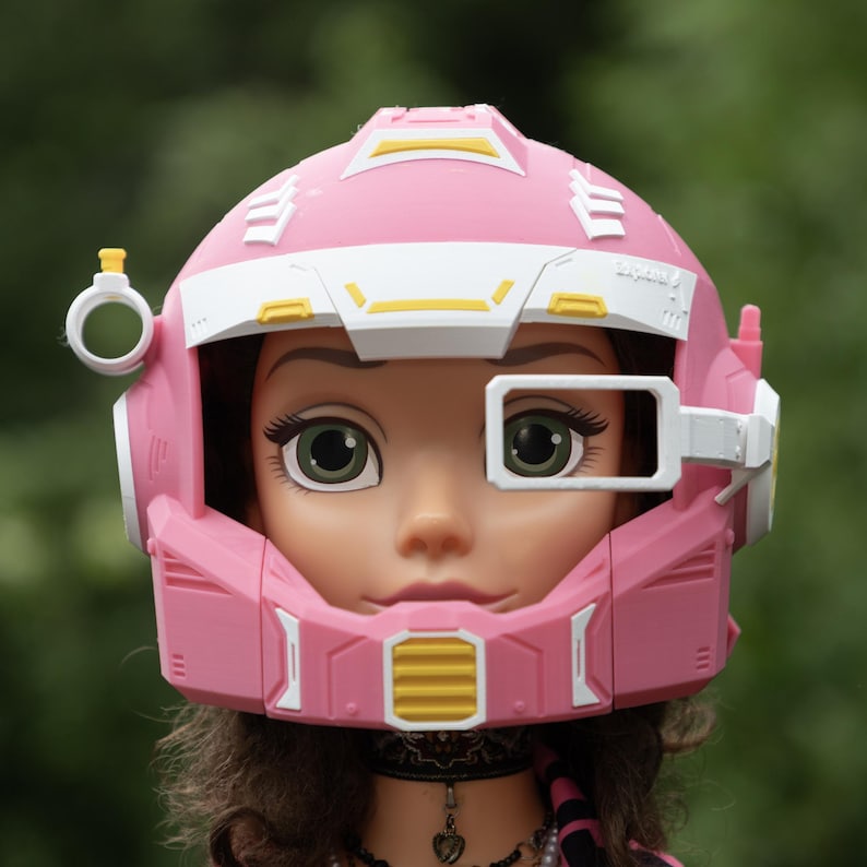 Pu&ograve; includere: Un casco futuristico rosa e bianco con accenti gialli indossato da una bambola. Il casco presenta un oculare rettangolare sopra l'occhio sinistro, un attacco circolare sul lato destro e un dettaglio di ventilazione giallo sulla mentoniera.