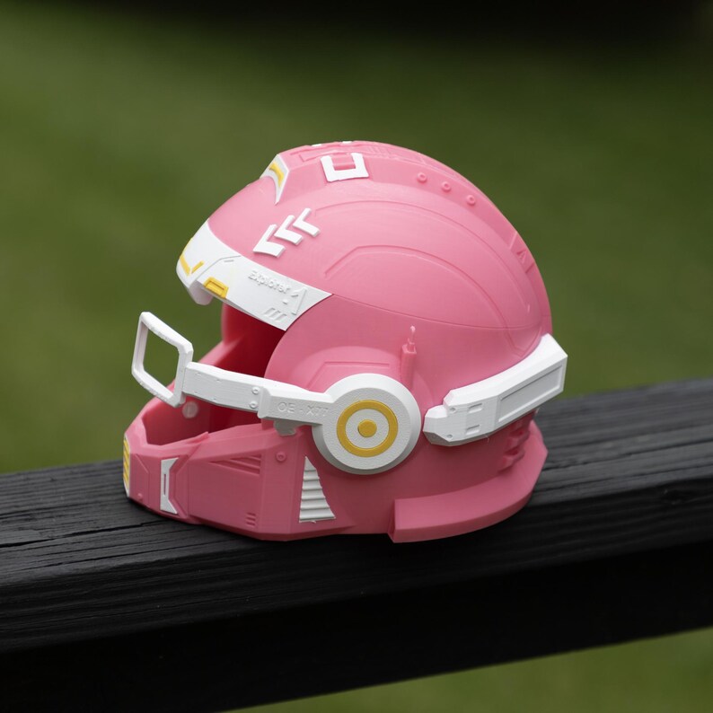 Pu&ograve; includere: Un casco rosa con accenti bianchi e gialli, inclusa una maschera facciale bianca, copriorecchie laterali circolari con centri gialli e dettagli geometrici bianchi sulla parte superiore. La parola Explorer &egrave; stampata in piccolo testo bianco sul lato.