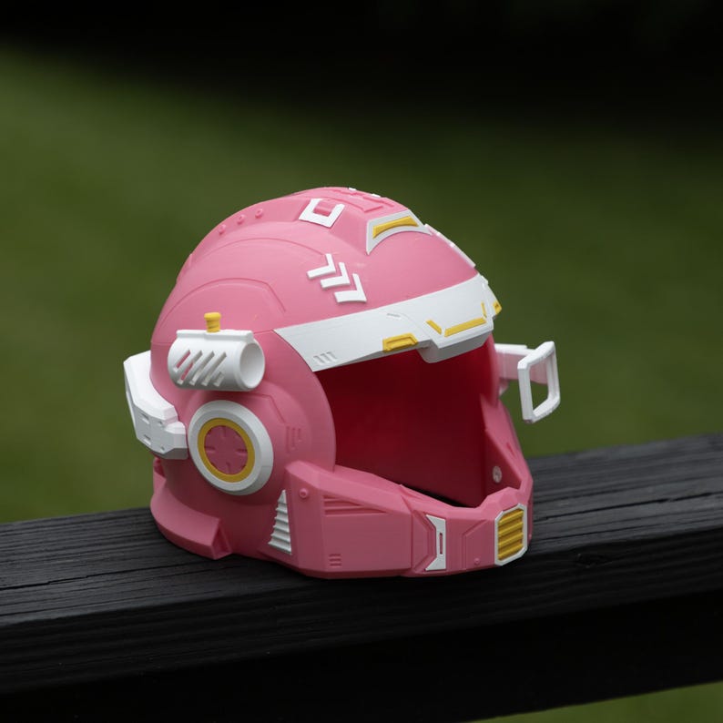 Pu&ograve; includere: Un casco rosa con accenti bianchi e gialli dal design futuristico. Il casco include un'area visiera bianca, attacchi laterali e dettagli geometrici modellati sulla calotta, creando un aspetto strutturato e tecnico.