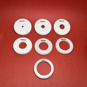 May include: Seven white circular objects with black text indicating sizes from 10mm to 70mm. Each object has a central hole, with the hole size increasing with the object's diameter. The objects are arranged on a red surface.