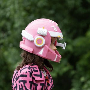 Pu&ograve; includere: Un casco in plastica rosa con accenti bianchi e gialli, dotato di un attacco rettangolare bianco laterale e dettagli geometrici stampati. Il casco &egrave; indossato sopra capelli castani e una giacca rosa con un motivo geometrico nero.