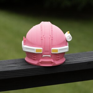 Pu&ograve; includere: Un casco rosa a forma di cupola con una fascia bianca sulla visiera e dettagli gialli. Il casco presenta una superficie modellata e strutturata con pannelli geometrici, una piccola antenna sul lato sinistro e un attacco cilindrico bianco a destra.