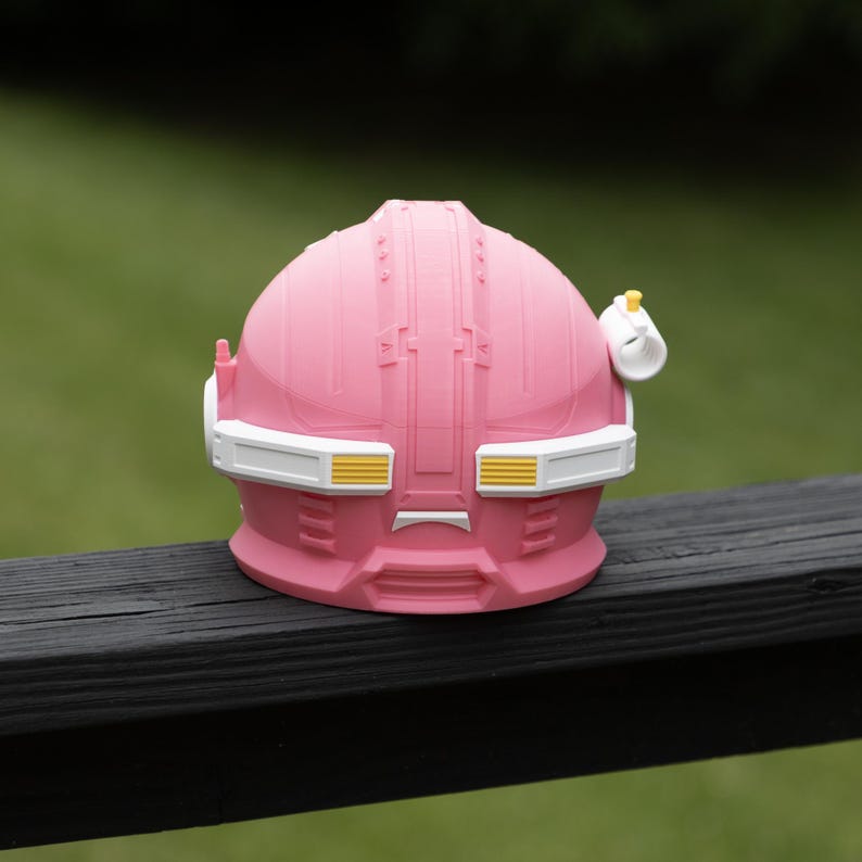 Pu&ograve; includere: Un casco rosa a forma di cupola con una fascia bianca sulla visiera e dettagli gialli. Il casco presenta una superficie modellata e strutturata con pannelli geometrici, una piccola antenna sul lato sinistro e un attacco cilindrico bianco a destra.