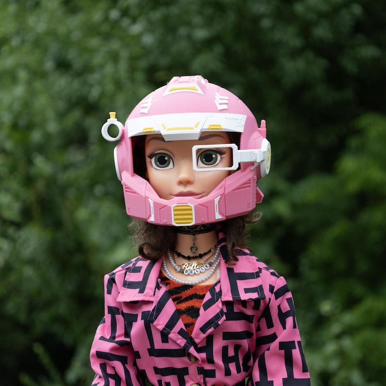 Pu&ograve; includere: Una bambola che indossa un casco futuristico rosa e bianco con un dettaglio di ventilazione giallo e un oculare bianco. La bambola ha i capelli castani e indossa una giacca rosa con un motivo geometrico nero sopra una camicia tigrata e collane.