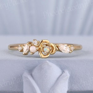 May include: A delicate gold ring with a floral design featuring a white opal gemstone and smaller clear gemstones. The ring is set on a light blue background.