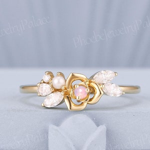 May include: A delicate gold ring featuring a rose design with a pink opal gemstone and white pearl accents. The ring is set with small clear gemstones and has a floral motif.