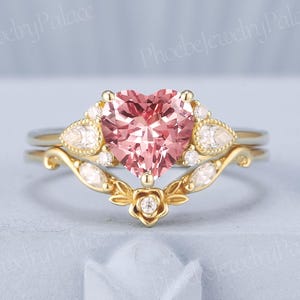 May include: A two-piece engagement ring set. The engagement ring features a heart-shaped pink gemstone, surrounded by smaller white gemstones. The band is gold-colored with floral and leaf designs. The wedding band is gold-colored with a curved design.