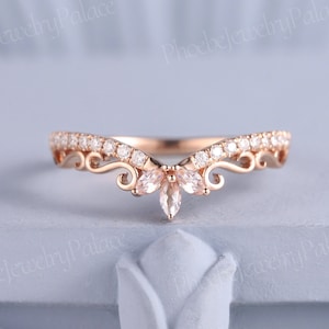 Op de afbeelding: Een delicate roségouden trouwring met een V-vormig ontwerp. De ring is versierd met kleine, glinsterende diamanten en een grotere, peervormige edelsteen in het midden.