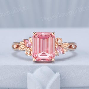 May include: A rose gold ring featuring a large, rectangular pink gemstone as the centerpiece. The ring is adorned with smaller pink gemstones and leaf-shaped accents on either side of the main stone, with small diamond accents.